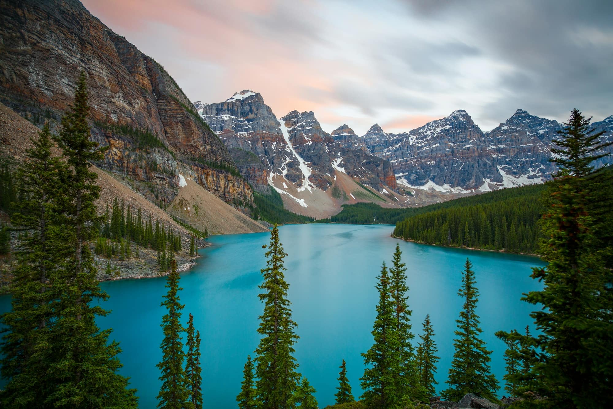 Snow-dusted mountain peaks above a turquoise lake, framed by tall spruce trees under a pink-streaked sky.