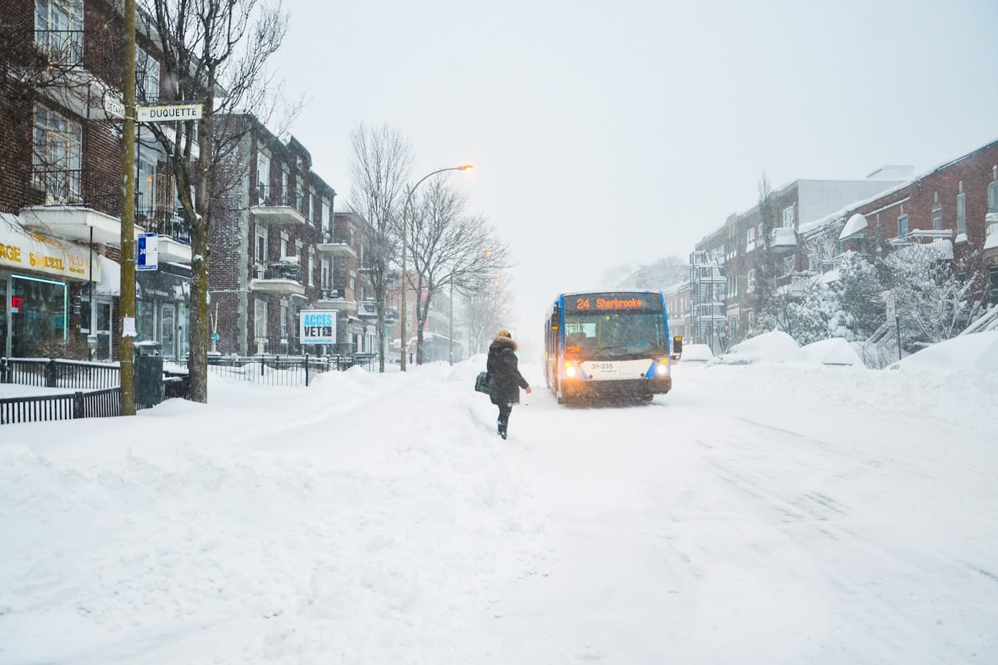 A lone pedestrian walking toward a bus through a snowstorm on a Montreal street.