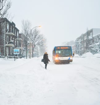 A lone pedestrian walking toward a bus through a snowstorm on a Montreal street.